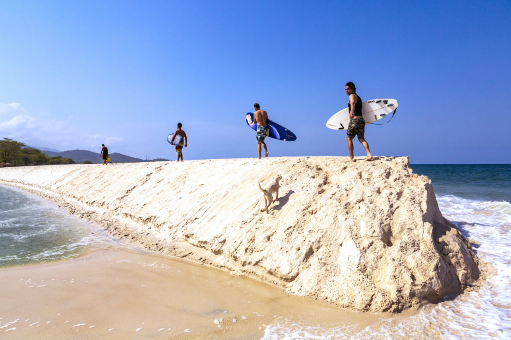 Vier Surfer laufen auf einem Sandrücken, den das Meer und ein Fluss in Sierra Leone gebildet haben, entlang.